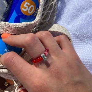 May include: A red and pink beaded ring with a silver rectangular charm. The ring is on a person's finger.