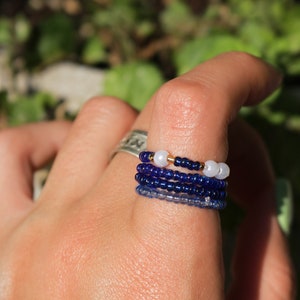 以下が含まれることがあります： 指に重ねられた4つのビーズリングのセット。リングは青と白のビーズで作られており、一番上のリングには金のビーズのアクセントが付けられています。