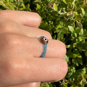 May include: A blue beaded ring with a brown evil eye charm.