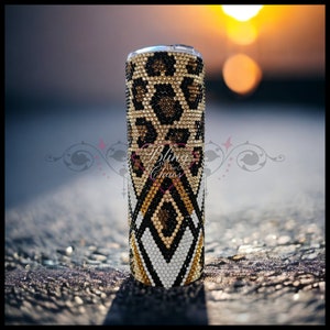 Op de afbeelding: Een met strass versierde beker met een luipaardprint aan de bovenkant en een geometrisch patroon aan de onderkant. De beker is bedekt met glinsterende strass in goud, zwart en wit. De achtergrond is wazig, met een zachte zonsondergang.