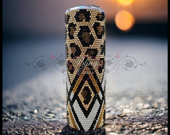 May include: A bedazzled tumbler with a leopard print design at the top and a geometric pattern at the bottom. The tumbler is covered in sparkling rhinestones in shades of gold, black, and white. The background is blurred, with a soft sunset.