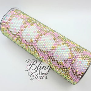 Puede incluir: Un vaso adornado con pedrería con tapa transparente. El vaso está cubierto de pedrería iridiscente en un patrón de rosa, blanco y dorado. El texto "Bling on the Chaos" es visible en la parte inferior del vaso.