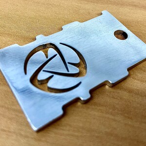 May include: A silver metal keychain with a geometric design and a hole for a ring. The keychain is made of brushed metal and has a smooth finish.