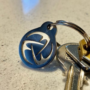 May include: A silver key ring with a circular charm featuring a stylised geometric design. The charm is attached to a silver ring and a set of keys.