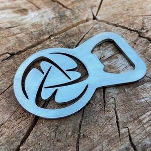 May include: A silver bottle opener with a circular design featuring a stylised, abstract pattern. The opener has a smooth, metallic finish.