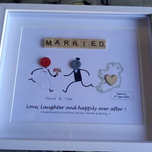 May include: A white frame with a white mount and a white background. The background has a hand-drawn illustration of a bride and groom running away from the altar. The bride is wearing a white dress and the groom is wearing a black suit. The word "MARRIED" is spelled out in wooden letter tiles. The date "9th April 2022" and the location "Tipperary" are written on the bottom right corner of the frame. The text "Love, Laughter and happily ever after! Congratulations love from Emma, Michael & family x" is written below the illustration.