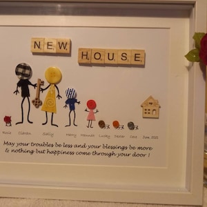 May include: A framed artwork with a message about a new house. The artwork features a family of stick figures made with buttons, a wooden house, and the word "NEW HOUSE" spelled out with wooden letter tiles. The message reads "May your troubles be less and your blessings be more & nothing but happiness come through your door!"