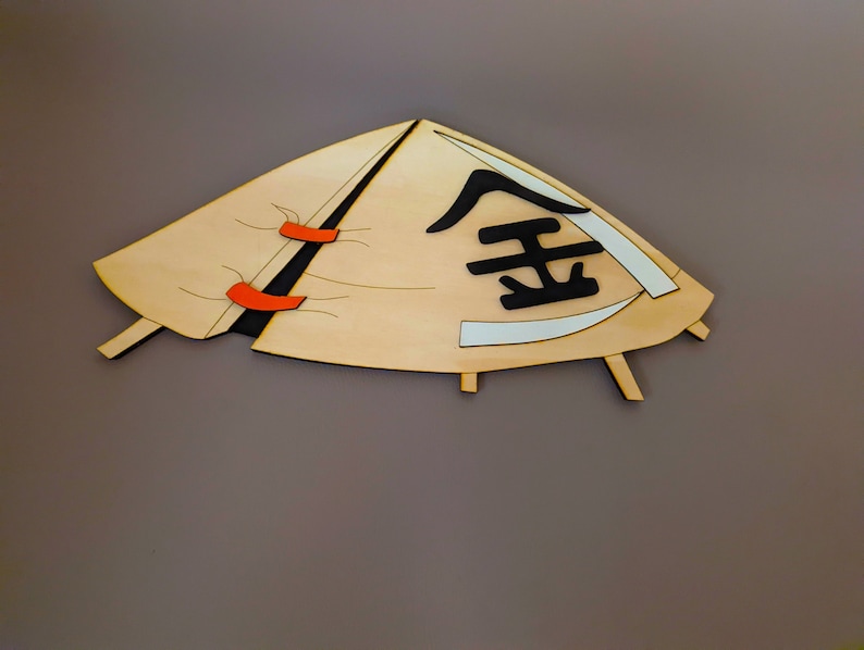 May include: A wooden cutout of a conical hat with a black kanji character on the front. The hat is beige with orange accents and is mounted on a gray background.