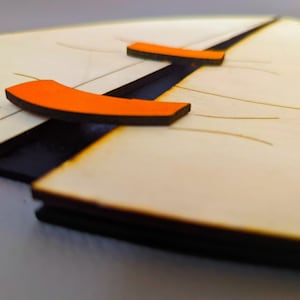 May include: A close-up of a wooden puzzle with two orange pieces. The pieces are shaped like a curved line and are attached to the puzzle with a black hinge.