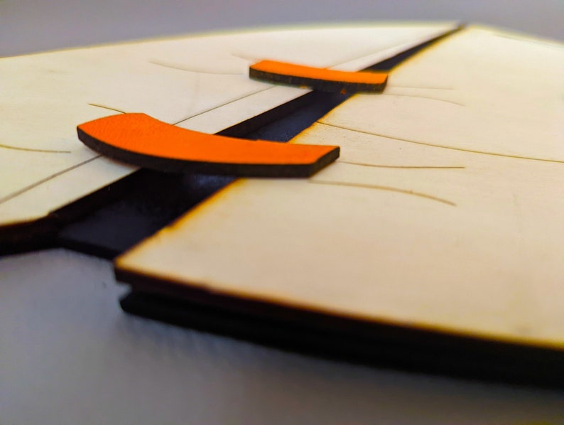 May include: A close-up of a wooden puzzle with two orange pieces. The pieces are shaped like a curved line and are attached to the puzzle with a black hinge.
