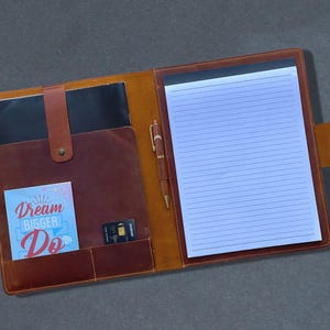 May include: Open leather portfolio with a notepad, pen, and card slots. The portfolio is brown with a lighter tan interior. A card with the text "Dream Bigger Do" is visible in a pocket.