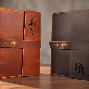 May include: Two leather journals with wrap-around closures and brass button studs. The left journal is tan with a mountain graphic and the text CHRISTOPHER HALL LEAD PLAN EXECUTE. The right journal is dark brown with the initials DR and DANIEL REED.