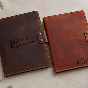 May include: Two rectangular leather notebook covers with snap closures. The dark brown cover is engraved with the name Peter Williams and a large letter P. The lighter brown cover is engraved with the name Joel Miller in a script font.