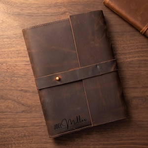 May include: A dark brown leather journal featuring a wrap-around strap secured with a single brass stud. The front cover is embossed with the name JOEL Miller in a black script font. The notebook has a smooth, matte finish and visible stitching.