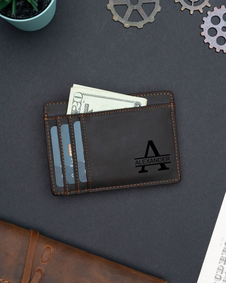 May include: A brown leather wallet with a personalized monogram of the letter "A" and the name "Alexander" in a black font. The wallet has a slot for cash and multiple card slots.