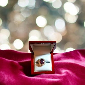 May include: A red velvet ring box with a gold ring featuring a large, oval-shaped ruby surrounded by smaller diamonds.