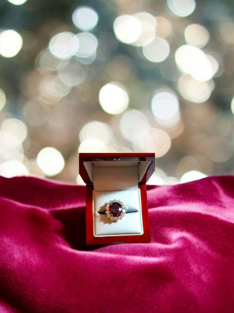 May include: A red velvet ring box with a gold ring featuring a large, oval-shaped ruby surrounded by smaller diamonds.