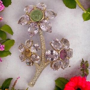 May include: A gold flower brooch with pink, yellow, and green gemstones. The center of the flower has a green stone with Hebrew writing.