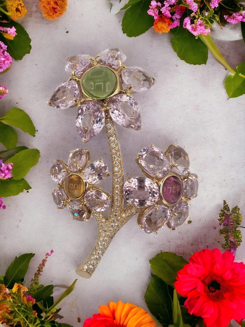 May include: A gold flower brooch with pink, yellow, and green gemstones. The center of the flower has a green stone with Hebrew writing.
