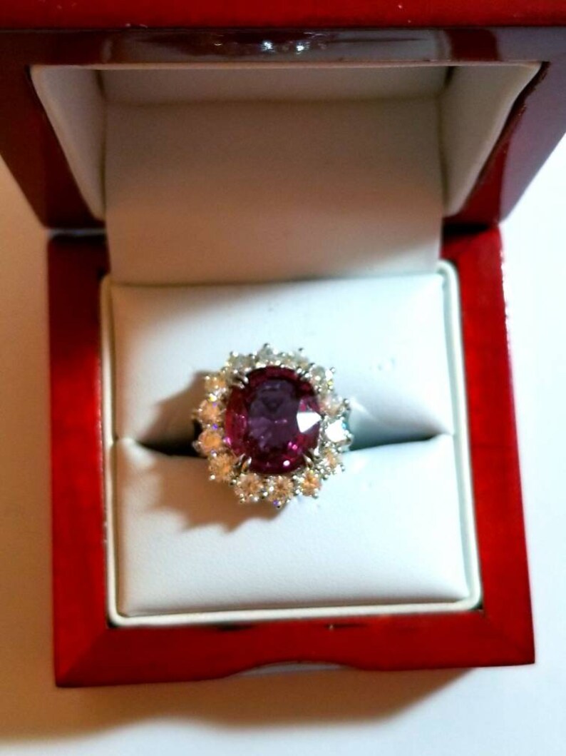 May include: A ring with a large oval-shaped pink gemstone surrounded by smaller diamonds in a white gold setting. The ring is in a red velvet ring box.
