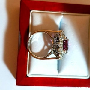 May include: A silver ring with a large, square-shaped, pink gemstone surrounded by smaller, clear gemstones. The ring is in a red velvet ring box.