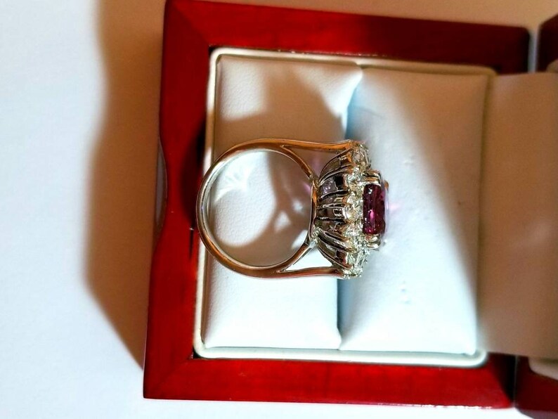 May include: A silver ring with a large, square-shaped, pink gemstone surrounded by smaller, clear gemstones. The ring is in a red velvet ring box.