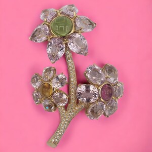 May include: A gold flower brooch with a green stone center and pink, yellow, and clear gemstones. The flower has a stem with small white gemstones.