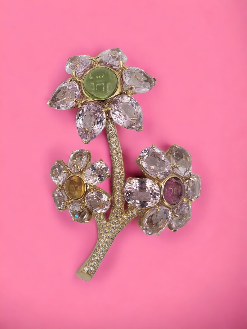 May include: A gold flower brooch with a green stone center and pink, yellow, and clear gemstones. The flower has a stem with small white gemstones.