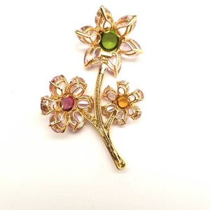 May include: A gold tone brooch with three flower designs. The flowers are made of pink and clear stones with a green, pink, and yellow center stone.
