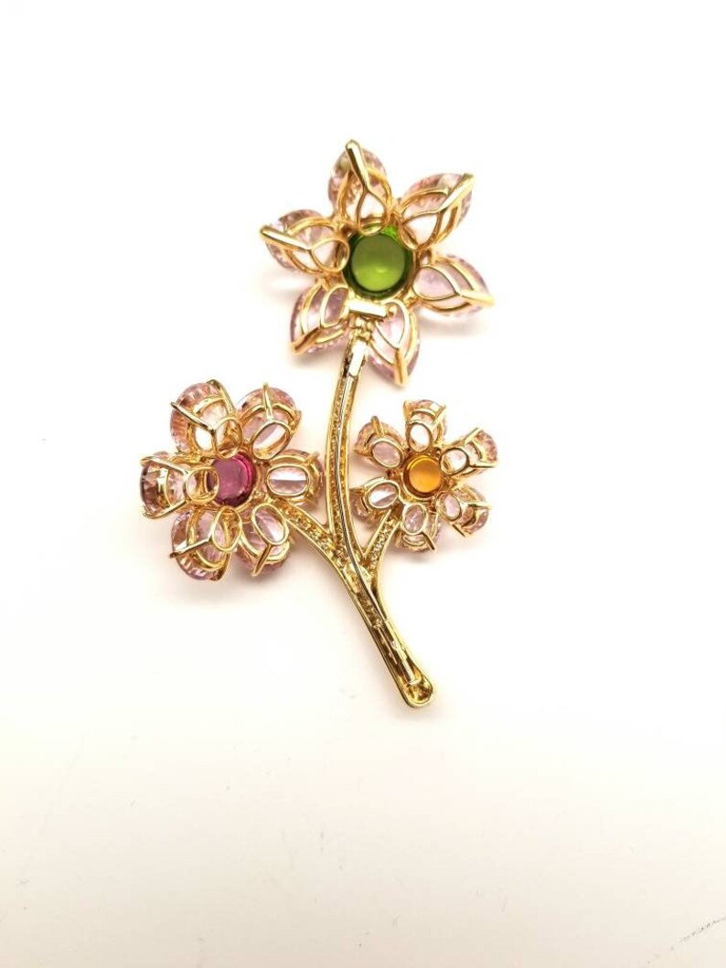 May include: A gold tone brooch with three flower designs. The flowers are made of pink and clear stones with a green, pink, and yellow center stone.