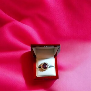 May include: A red gemstone ring with a halo of smaller diamonds set in a gold band. The ring is displayed in a red velvet ring box.