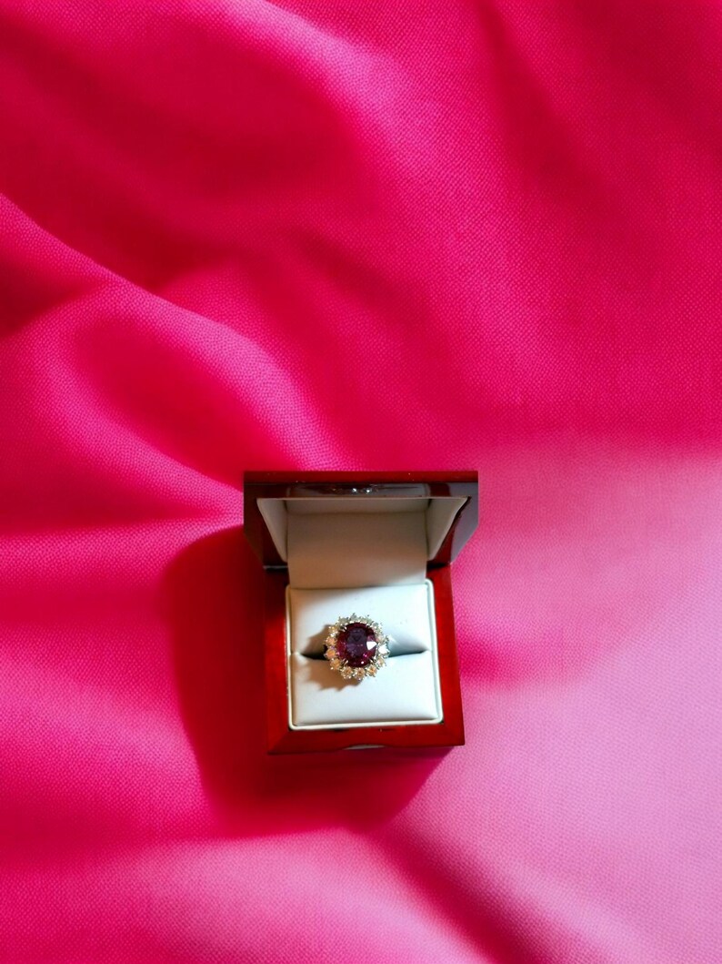 May include: A red gemstone ring with a halo of smaller diamonds set in a gold band. The ring is displayed in a red velvet ring box.