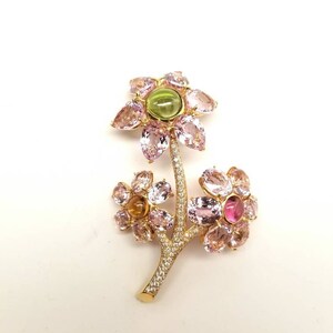 May include: A gold brooch in the shape of a flower with pink, green, and yellow gemstones. The brooch has a delicate stem with small diamonds.