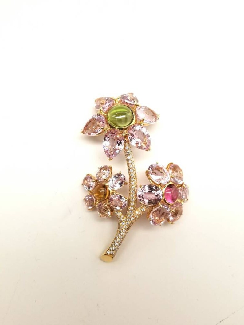 May include: A gold brooch in the shape of a flower with pink, green, and yellow gemstones. The brooch has a delicate stem with small diamonds.