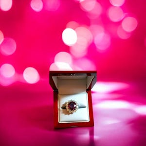 May include: A red velvet ring box with a large, oval-shaped ruby ring with diamond accents. The ring is in focus and the box is slightly out of focus. The background is a pink bokeh.