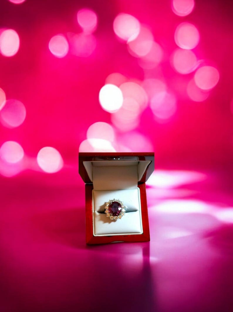 May include: A red velvet ring box with a large, oval-shaped ruby ring with diamond accents. The ring is in focus and the box is slightly out of focus. The background is a pink bokeh.