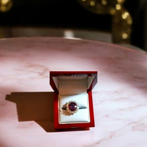 May include: A red ring box with a gold ring inside. The ring has a large, oval-shaped, dark red gemstone surrounded by smaller, white gemstones.
