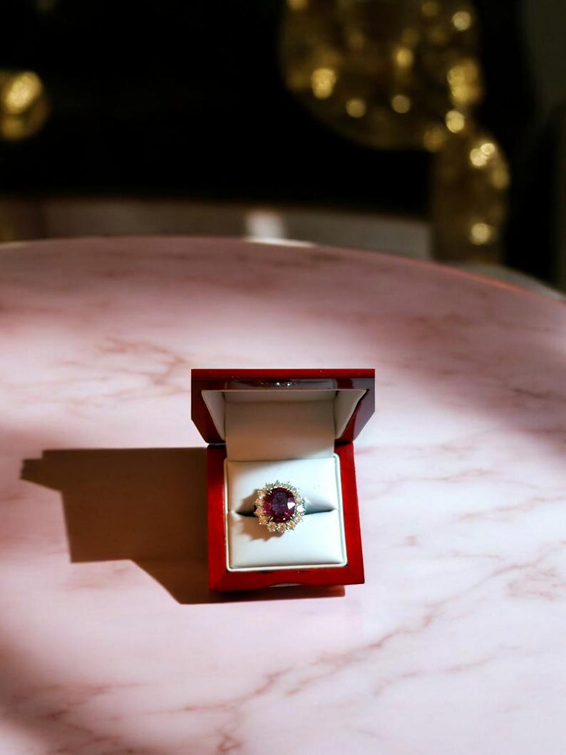 May include: A red ring box with a gold ring inside. The ring has a large, oval-shaped, dark red gemstone surrounded by smaller, white gemstones.