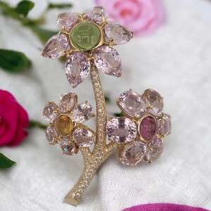 May include: A gold brooch in the shape of a flower with pink and green gemstones. The center of the flower has a green stone with the word "Jerusalem" engraved on it.