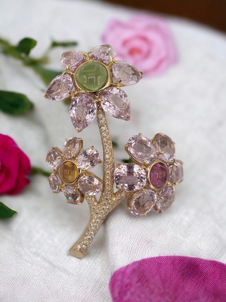 May include: A gold brooch in the shape of a flower with pink and green gemstones. The center of the flower has a green stone with the word "Jerusalem" engraved on it.