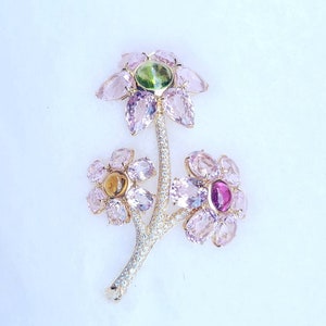 May include: A gold brooch with three pink flowers, each with a different colored gemstone in the center. The flowers are connected by a gold stem with a diamond-like design.