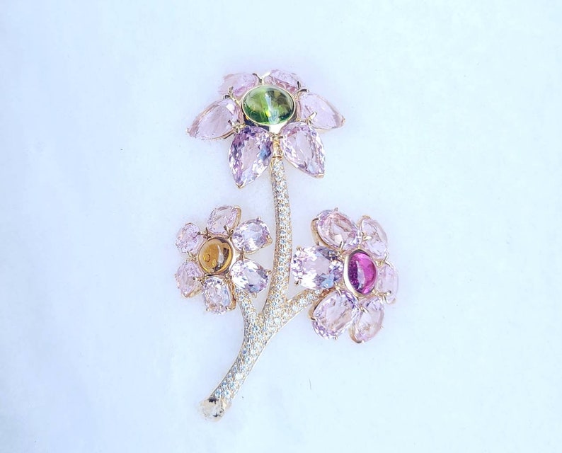 May include: A gold brooch with three pink flowers, each with a different colored gemstone in the center. The flowers are connected by a gold stem with a diamond-like design.