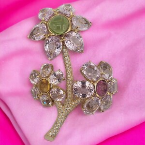 May include: A gold flower brooch with a green stone in the center of the top flower. The brooch is made of clear and pink stones and has a diamond-encrusted stem.