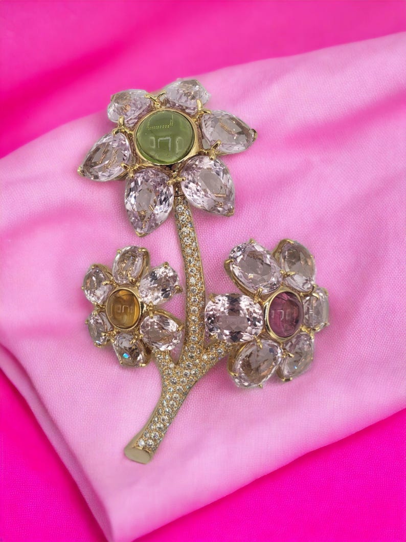 May include: A gold flower brooch with a green stone in the center of the top flower. The brooch is made of clear and pink stones and has a diamond-encrusted stem.
