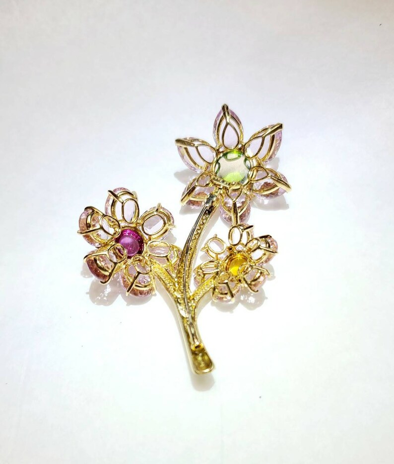 May include: A gold brooch with three flowers made of pink, green, and yellow gemstones. The flowers are connected by a gold stem.