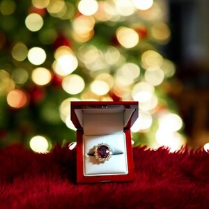 May include: A red velvet ring box with a sparkling ruby ring with diamond accents. The ring is in focus and the box is open. The background is a blurry image of a Christmas tree with lights.