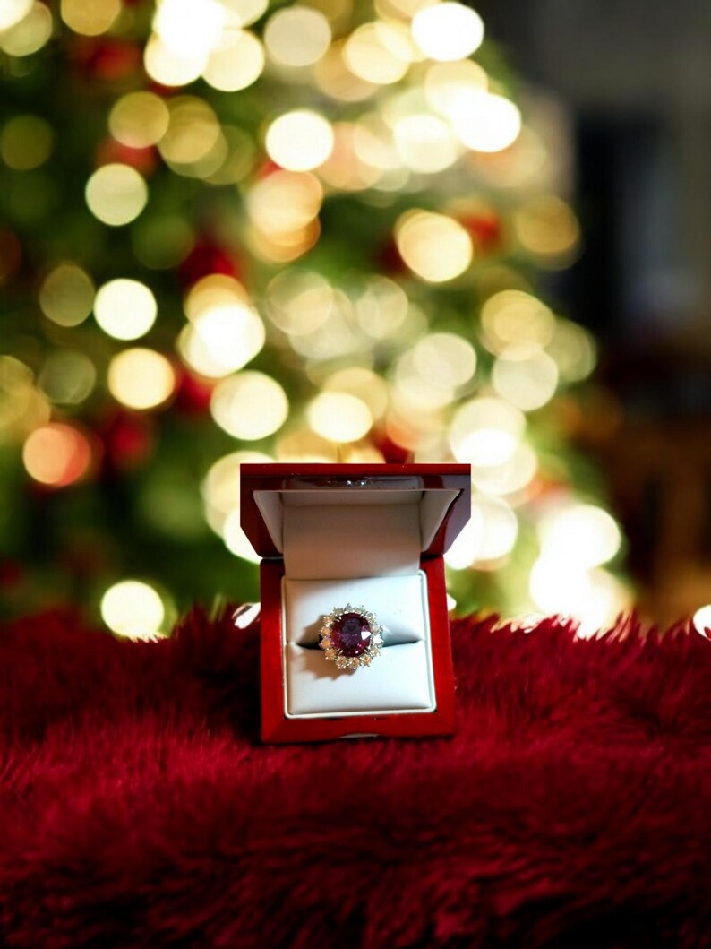 May include: A red velvet ring box with a sparkling ruby ring with diamond accents. The ring is in focus and the box is open. The background is a blurry image of a Christmas tree with lights.