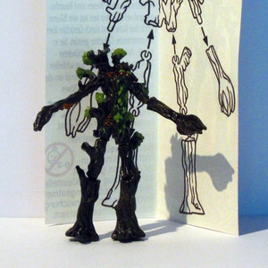 May include: A green and brown plastic toy tree creature with a face. The toy is assembled from multiple pieces, and the instructions for assembly are shown on a white sheet of paper behind the toy.