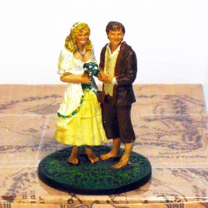 Puede incluir: Una figura en miniatura de una pareja sobre una base circular verde, colocada sobre una caja de cartón. La caja tiene una etiqueta con el texto "Lord of the Rings", "Sam & Rosie wedding at Hobbiton" y "Nr: 54.K". Las figuras están descalzas.