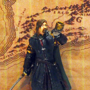 May include: A hand-painted miniature figurine of a warrior wearing a dark blue coat and cloak, holding a sword in the right hand and blowing into a horn held to the mouth with the left hand. The figure stands on a circular black base.